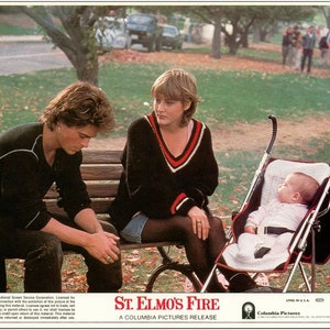 May include: A scene from the film "St. Elmo's Fire" with three people sitting on a park bench. A woman in a black sweater with a red stripe is sitting on the bench with a baby in a pushchair next to her. A man in a black shirt is sitting on the bench next to her. The film title "St. Elmo's Fire" is displayed at the bottom of the image.