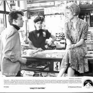May include: Black and white promotional still from the film "Pretty in Pink." A man in a blazer and turtleneck speaks to two women. One woman sits in a patterned dress, and the other stands behind a counter. The film's title is at the bottom.