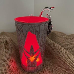 May include: A cylindrical cup with a textured gray exterior resembling tree bark and a bright red interior. A cutout in the shape of a flame glows with red light on the front. A small white, black, and red dragon figure is attached to the rim.