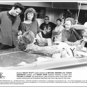 Puede incluir: Una fotografía en blanco y negro de un set de filmación con actores y equipo. Los actores están de pie alrededor de un automóvil, y el director los observa ensayando una escena. El texto en la foto dice: "El director Ridley Scott (derecha) observa a Michael Madsen (izquierda), Susan Sarandon (centro) y Geena Davis (abajo) ensayando una escena de Thelma & Louise, la historia aventurera y a menudo humorística de dos mujeres trabajadoras cuyas frustraciones reprimidas las llevan a una carrera de cuatro días por sus vidas."