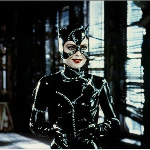 Könnte beinhalten: Eine Frau in einem schwarzen Leder-Catsuit mit einer schwarzen Maske und Katzenohren. Sie steht in einem Raum mit einem Fenster hinter ihr.