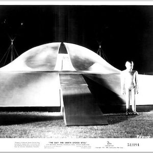 以下が含まれることがあります： 1951年のSF映画「地球が静止する日」のシーンを捉えた白黒写真。前景には円盤型の巨大な宇宙船が映っており、その前に人間のような宇宙人が立っている。