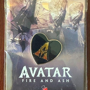 May include: A heart-shaped enamel pin featuring a gold-colored letter A on a black background, displayed on a promotional card for the film Avatar: Fire and Ash, which includes the movie title and the Variety logo in the lower section.