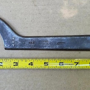 May include: A dark grey metal hook spanner wrench with a curved end and a small protruding pin. The tool features the stamped number 464 and is positioned above a yellow measuring tape, showing a total length of approximately 10 inches.