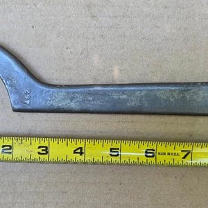 May include: A gray metal hook spanner wrench with a curved end and a small cylindrical pin, featuring the number 297 stamped on the side. A yellow measuring tape is positioned below the tool, indicating a total length of approximately 9 inches.