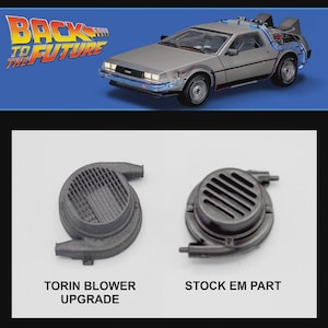 Puede incluir: Un modelo de coche DeLorean plateado con detalles azules, inspirado en la película "Regreso al Futuro", se muestra sobre dos piezas de automóvil negras. Una pieza está etiquetada como "TORIN BLOWER UPGRADE", la otra como "STOCK EM PART".