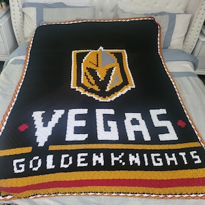May include: A black crocheted blanket featuring a gold and white Vegas Golden Knights logo. The logo depicts a knight's helmet with a sword and shield.