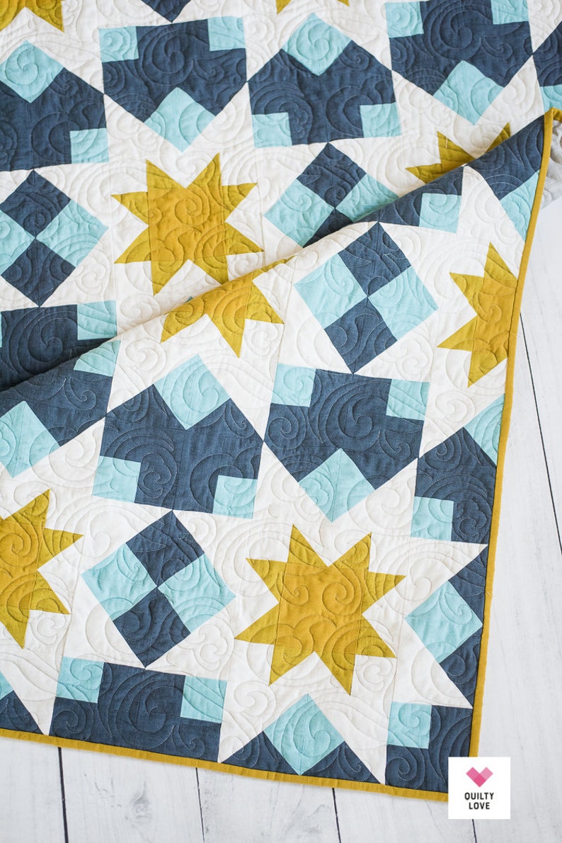 May include: A quilt with a geometric pattern of blue, white and yellow stars. The quilt is folded in half and the fabric has a quilted texture.