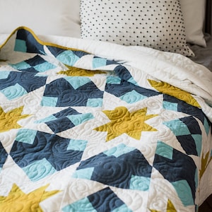 May include: A white, blue and gold quilt with a star pattern. The quilt is folded on a bed with a white pillow and a black and white patterned pillow. The quilt has a "Quilty Love" logo in the bottom right corner.