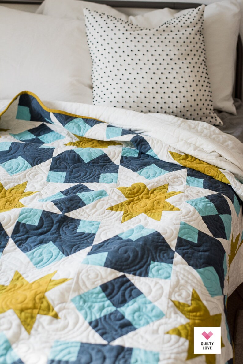 May include: A white, blue and gold quilt with a star pattern. The quilt is folded on a bed with a white pillow and a black and white patterned pillow. The quilt has a "Quilty Love" logo in the bottom right corner.