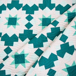May include: A teal and white quilt with a geometric pattern of stars and squares. The quilt is folded in half, revealing the intricate stitching.