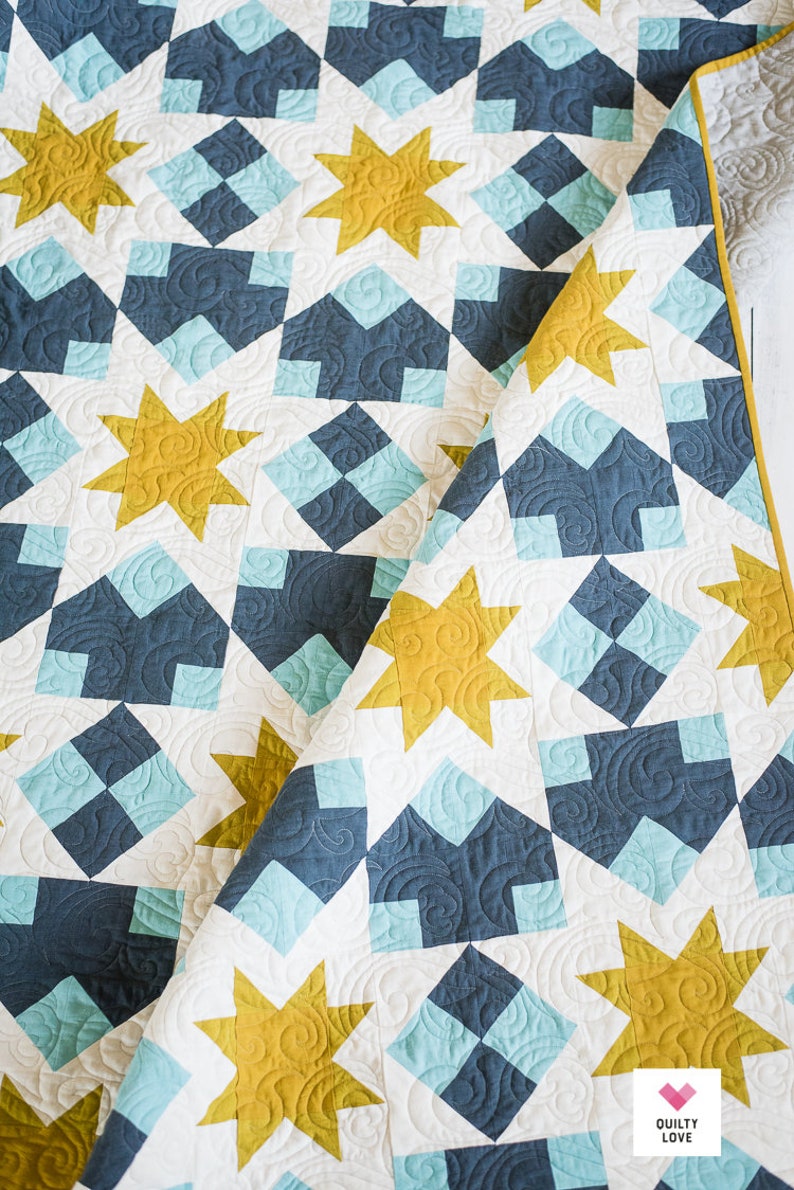May include: A quilt with a white background and a pattern of blue and teal squares and yellow stars. The quilt is folded in half and the text "Quilty Love" is visible in the bottom right corner.