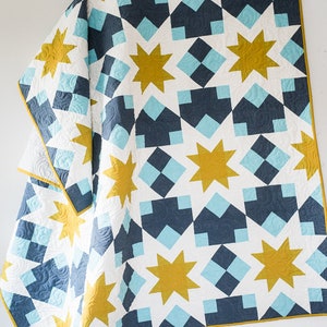 May include: A white, blue and gold quilt with a geometric pattern of stars and squares. The quilt is folded in half and draped over a white surface.