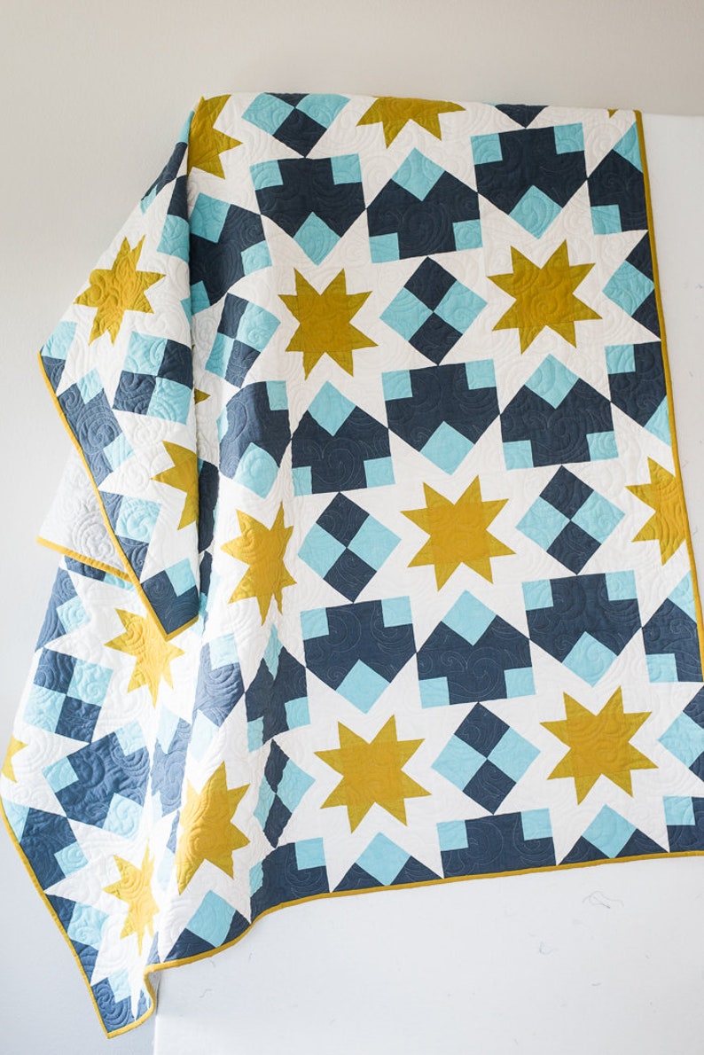 May include: A white, blue and gold quilt with a geometric pattern of stars and squares. The quilt is folded in half and draped over a white surface.