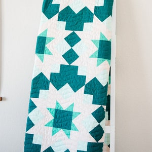 May include: A teal and white quilt with a geometric pattern. The quilt is folded and hanging on a white ladder.
