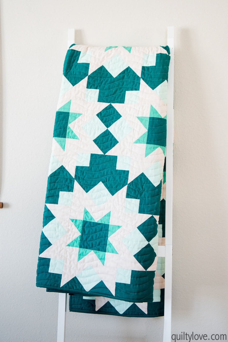 May include: A teal and white quilt with a geometric pattern. The quilt is folded and hanging on a white ladder.