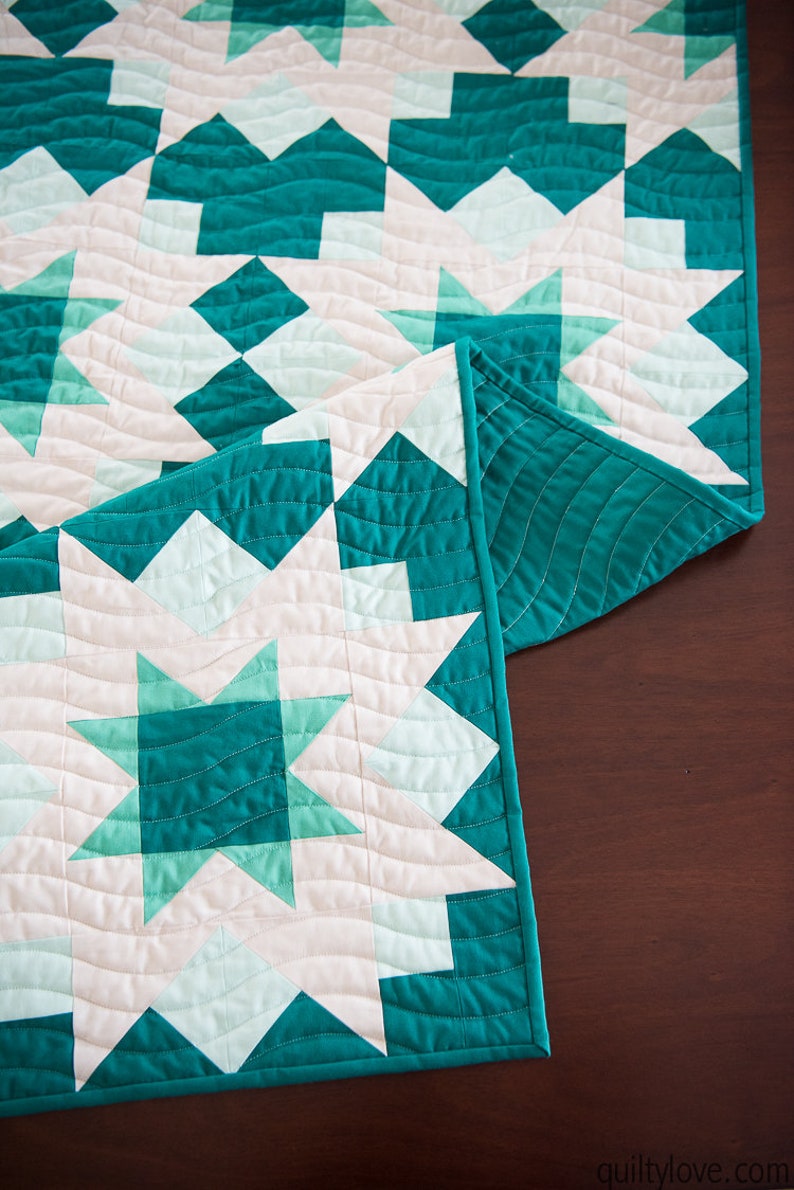 May include: A teal and white quilted table runner with a star pattern. The runner is folded in half and is lying on a wooden surface.
