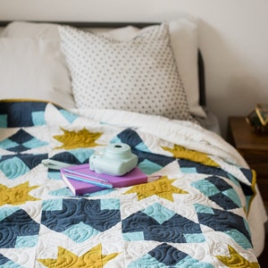May include: A white, blue and gold quilt with a star pattern. A purple notebook and a blue camera are on top of the quilt.