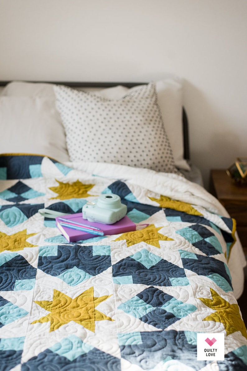 May include: A white, blue and gold quilt with a star pattern. A purple notebook and a blue camera are on top of the quilt.