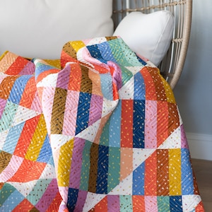 May include: A colorful patchwork quilt with a geometric pattern of stripes and triangles. The quilt is made with a variety of fabrics in shades of orange, pink, blue, green, and brown. The quilt is folded and draped over a white wicker chair. The quilt has a yellow border and a small logo that says "Quilty Love" in the bottom right corner.