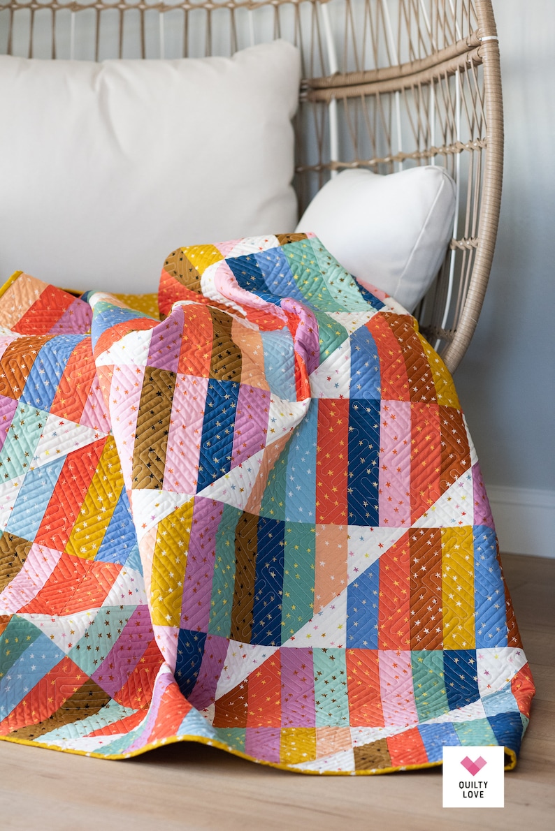 May include: A colorful patchwork quilt with a geometric pattern of stripes and triangles. The quilt is made with a variety of fabrics in shades of orange, pink, blue, green, and brown. The quilt is folded and draped over a white wicker chair. The quilt has a yellow border and a small logo that says "Quilty Love" in the bottom right corner.