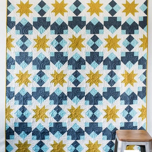 May include: A quilt with a geometric pattern of white, blue and gold stars. The quilt is hanging on a white wall with a wooden stool in the foreground.