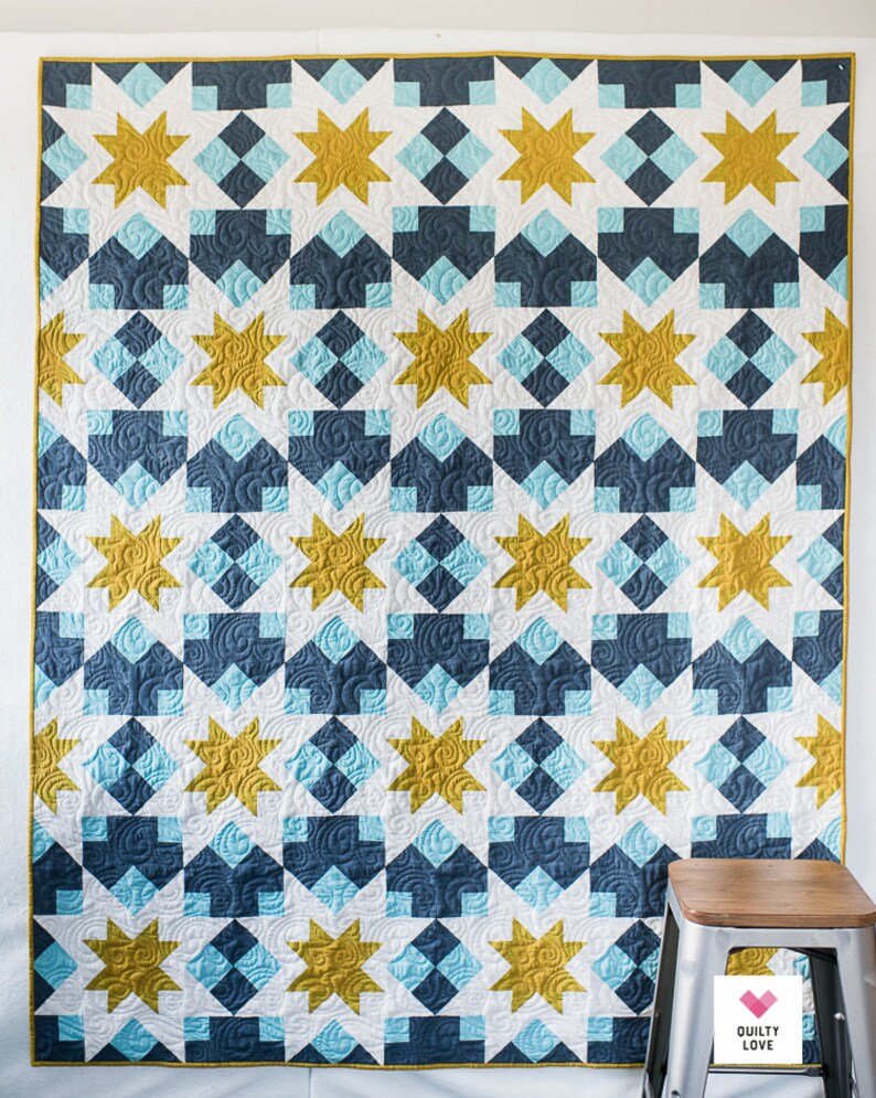 May include: A quilt with a geometric pattern of white, blue and gold stars. The quilt is hanging on a white wall with a wooden stool in the foreground.