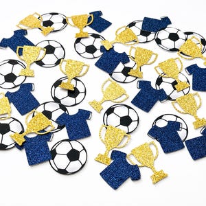 May include: A selection of football-themed confetti, featuring black and white footballs, gold glitter trophies, and navy blue glitter t-shirts. The confetti is spread out on a white background, creating a celebratory feel.