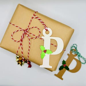 May include: A brown paper gift bag tied with red and white twine. A white paper gift tag with the letter "P" and a green holly sprig is attached to the bag. A second brown paper gift tag with the letter "P" and a green holly sprig is on the table.