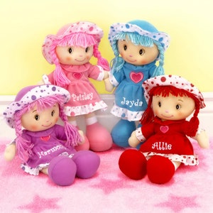 Peut inclure: Quatre poup&eacute;es de chiffon faites &agrave; la main, v&ecirc;tues de robes roses, bleues, violettes et rouges, chacune avec un chapeau assorti. Les poup&eacute;es ont des visages brod&eacute;s, des cheveux en fil et les noms "Paisley", "Jayda", "Harmony" et "Allie" brod&eacute;s sur leurs robes. Elles sont assises sur un tapis rose &agrave; motifs d'&eacute;toiles.