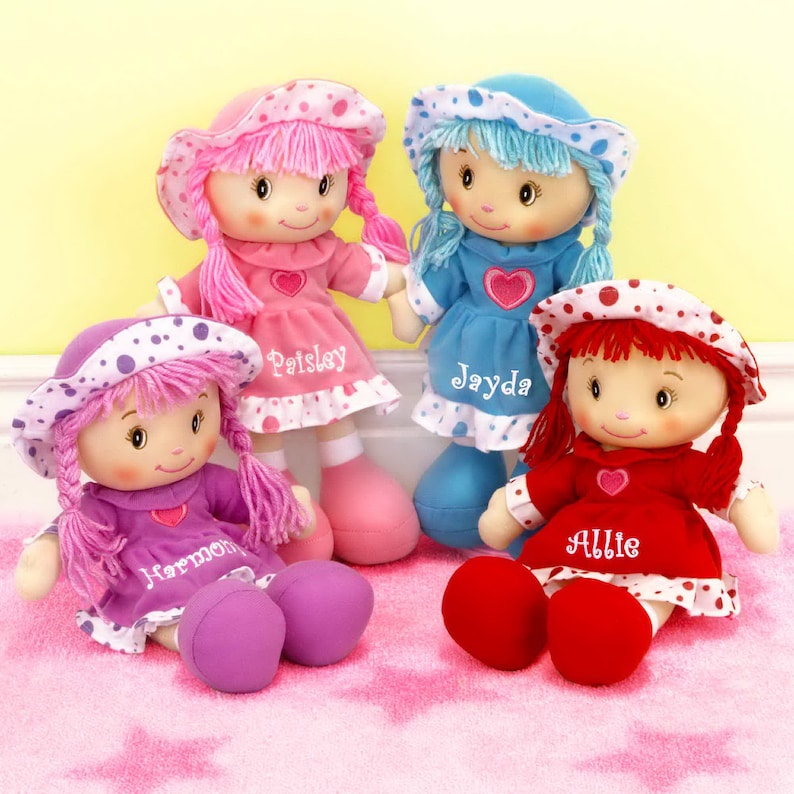 Peut inclure: Quatre poup&eacute;es de chiffon faites &agrave; la main, v&ecirc;tues de robes roses, bleues, violettes et rouges, chacune avec un chapeau assorti. Les poup&eacute;es ont des visages brod&eacute;s, des cheveux en fil et les noms "Paisley", "Jayda", "Harmony" et "Allie" brod&eacute;s sur leurs robes. Elles sont assises sur un tapis rose &agrave; motifs d'&eacute;toiles.