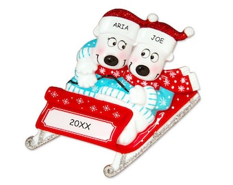 May include: A personalized Christmas ornament featuring two polar bears wearing blue and white sweaters and red hats, riding a red sled with a silver trim. The sled has a nameplate that reads "20XX". The bears are wearing hats that say "ARIA" and "JOE".