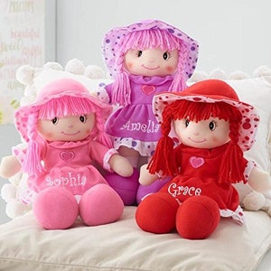Peut inclure: Trois poup&eacute;es en peluche avec des chapeaux et des robes roses et rouges. Les poup&eacute;es portent les noms brod&eacute;s : Sophia, Amelia et Grace. Chaque poup&eacute;e a un c&oelig;ur et un visage souriant.