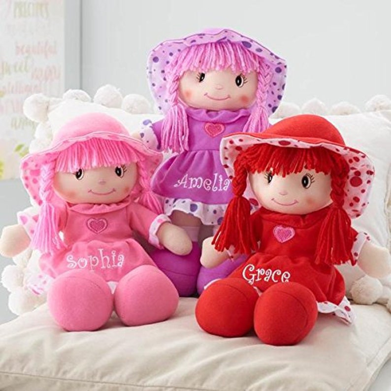 Peut inclure: Trois poup&eacute;es en peluche avec des chapeaux et des robes roses et rouges. Les poup&eacute;es portent les noms brod&eacute;s : Sophia, Amelia et Grace. Chaque poup&eacute;e a un c&oelig;ur et un visage souriant.