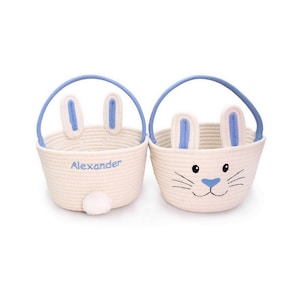 May include: Two cream-colored Easter baskets with blue accents. One basket has the name "Alexander" embroidered in blue. Both baskets feature bunny ears and handles, with a bunny face design on one.