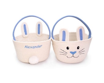 May include: Two cream-colored Easter baskets with blue accents. One basket has the name "Alexander" embroidered in blue. Both baskets feature bunny ears and handles, with a bunny face design on one.