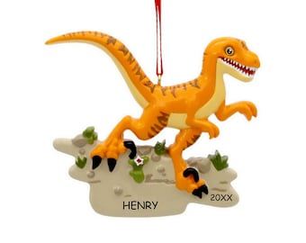 May include: An orange dinosaur ornament with brown stripes, standing on a beige rocky base with green accents. The base features the text HENRY and 20XX. A thin red ribbon is attached to the top for hanging, and the dinosaur has sharp teeth.