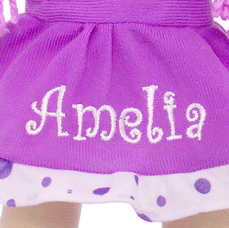 Peut inclure: Gros plan sur une jupe de poup&eacute;e violette brod&eacute;e du nom "Amelia" en &eacute;criture blanche. La jupe est orn&eacute;e d'une bordure &agrave; pois blancs. La tenue de la poup&eacute;e est en tissu doux.