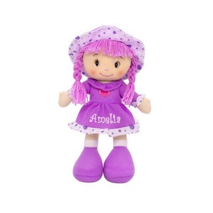 Peut inclure: Une poup&eacute;e en peluche douce avec une robe violette et un chapeau assorti. La poup&eacute;e a des cheveux roses en fil tress&eacute;s et le nom "Amelia" brod&eacute; sur la robe. Le chapeau et la robe sont d&eacute;cor&eacute;s de pois blancs. La poup&eacute;e a un c&oelig;ur sur la poitrine.