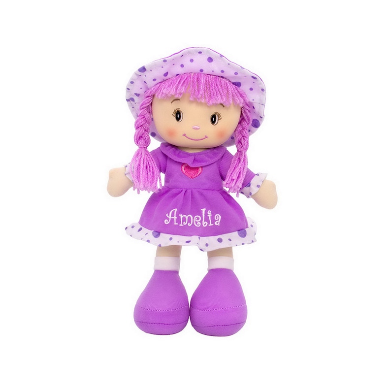 Peut inclure: Une poup&eacute;e en peluche douce avec une robe violette et un chapeau assorti. La poup&eacute;e a des cheveux roses en fil tress&eacute;s et le nom "Amelia" brod&eacute; sur la robe. Le chapeau et la robe sont d&eacute;cor&eacute;s de pois blancs. La poup&eacute;e a un c&oelig;ur sur la poitrine.