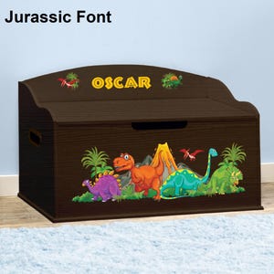 May include: A brown wooden toy chest with a dinosaur theme. The chest has a hinged lid and a personalized nameplate that reads "OSCAR". The front of the chest features a colorful illustration of dinosaurs, a volcano, and palm trees.