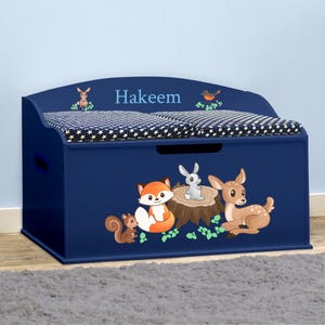 May include: A blue wooden toy chest with a padded lid and a woodland animal theme. The lid has a star pattern and the name "Hakeem" is written on the front. The chest features a fox, a deer, a squirrel, and a rabbit.