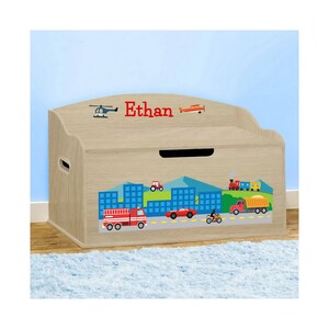 May include: A light brown wooden toy chest with a city scene design. The chest features a red name "Ethan" and illustrations of vehicles, including a helicopter, airplane, fire truck, and cars. The chest has a curved top and a handle cutout.