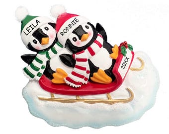 Puede incluir: Un adorno navideño personalizado con dos pingüinos que llevan gorros y bufandas, montando un trineo rojo sobre un fondo de nube blanca. Los pingüinos se llaman "Leila" y "Ronnie" y el año "20XX" está escrito en el trineo.