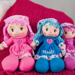 Peut inclure: Trois poup&eacute;es de chiffon en peluche avec des visages brod&eacute;s, chacune portant une robe et un bonnet. Les poup&eacute;es ont des cheveux roses, bleus et violets, et leurs robes sont de couleurs assorties. Chaque poup&eacute;e a un nom brod&eacute; sur la robe.