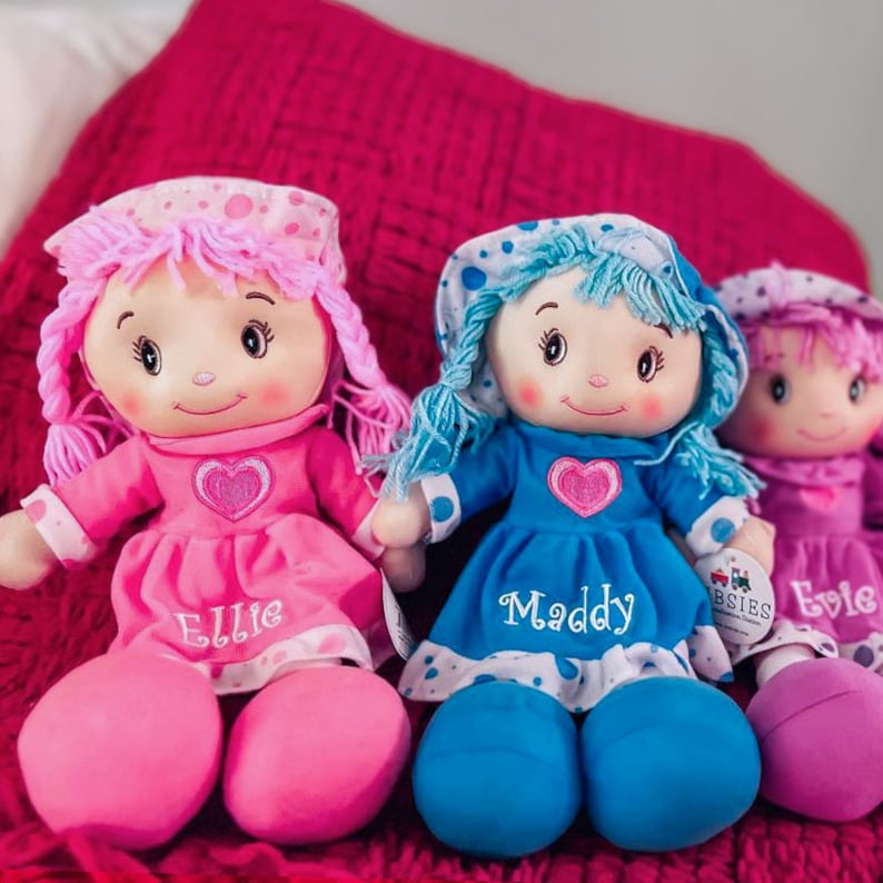 Peut inclure: Trois poup&eacute;es de chiffon en peluche avec des visages brod&eacute;s, chacune portant une robe et un bonnet. Les poup&eacute;es ont des cheveux roses, bleus et violets, et leurs robes sont de couleurs assorties. Chaque poup&eacute;e a un nom brod&eacute; sur la robe.