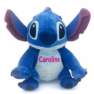 May include: A plush blue character with large ears and a white belly, embroidered with the name "Caroline" in pink. The character has large black eyes and is a popular children's toy.
