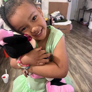 May include: A young girl wearing a green dress with a pink and black stuffed animal. She is smiling and holding the stuffed animal close to her.