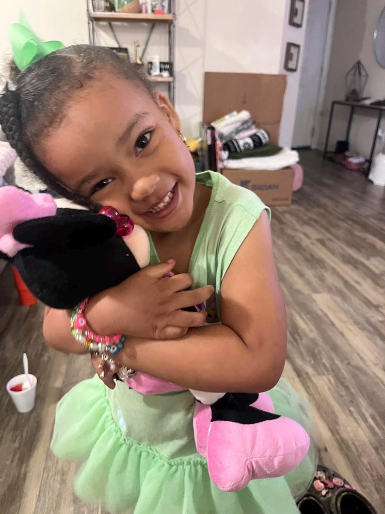 May include: A young girl wearing a green dress with a pink and black stuffed animal. She is smiling and holding the stuffed animal close to her.