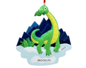 May include: A green long-necked dinosaur ornament with a yellow underbelly, holding a green leaf in its mouth. It stands on a light blue snowy base with the text BROOKLYN and 20XX, set against a backdrop of dark blue jagged mountains.